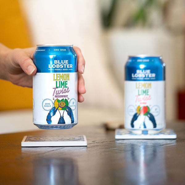Person holding a can of Blue Lobster Lemon Lime Passionfruit Twist drink with another can on a coaster in the background.